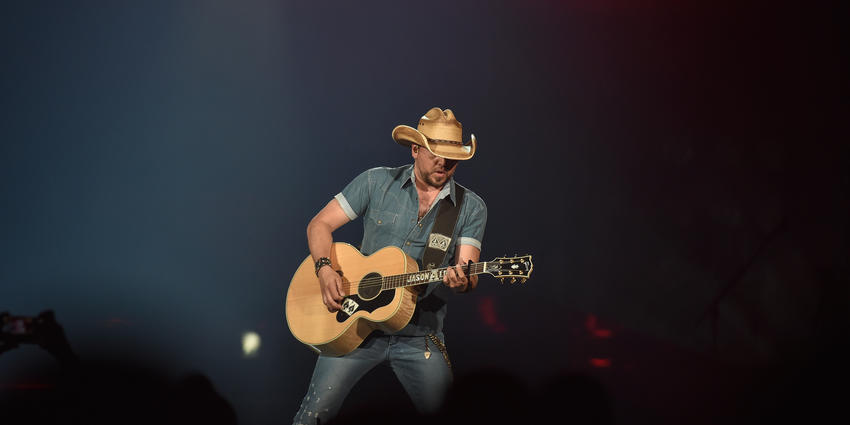 Jason Aldean, playing guitar, wearing his signature cowboy hat and short sleeved western buttoned down western shirt.